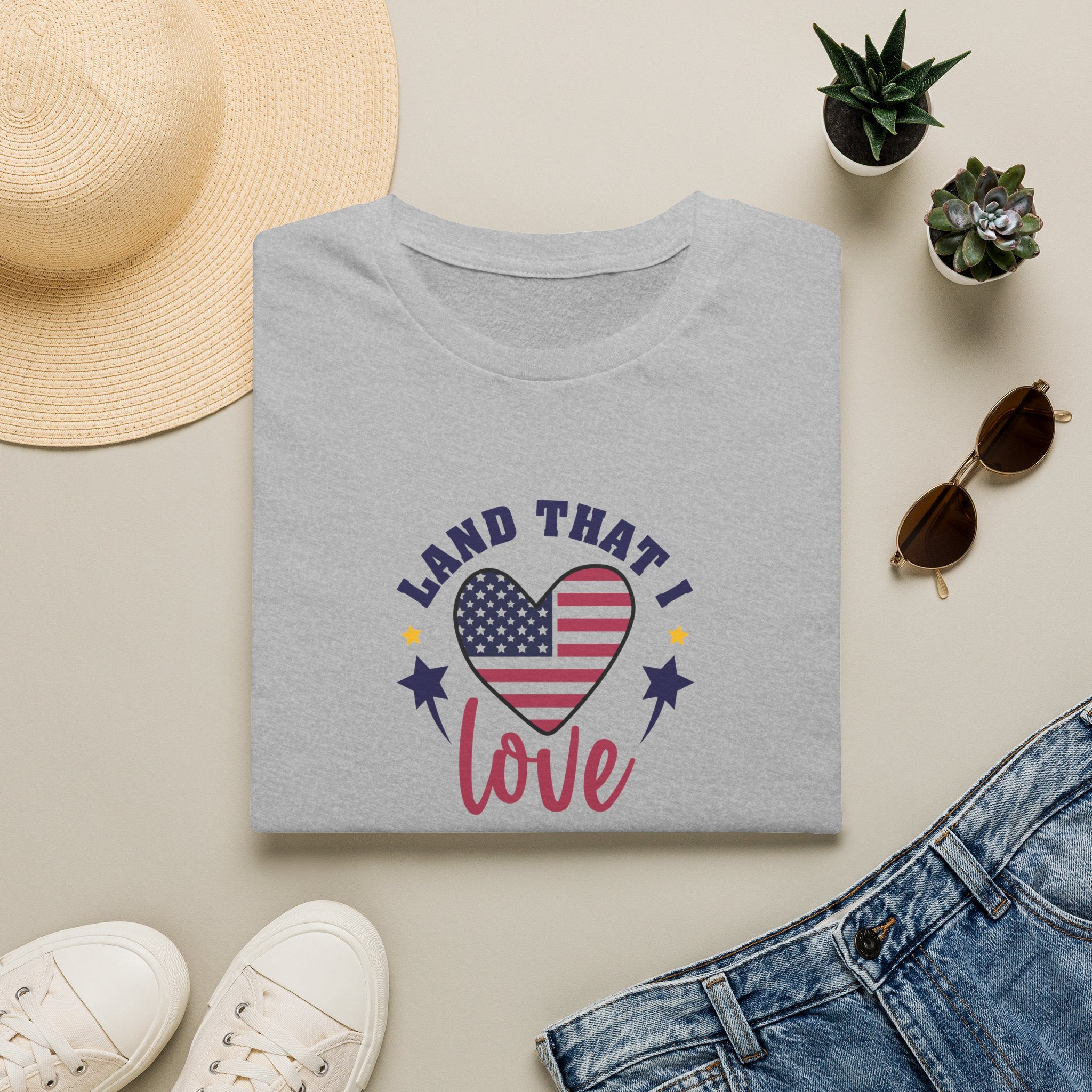 Light gray patriotic t-shirt with American flag heart design saying "Land That I Love" on a flat lay background.